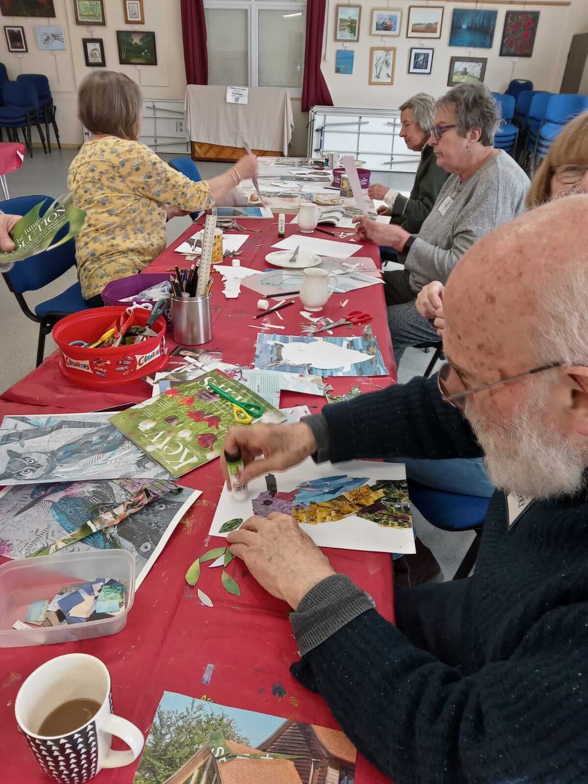There is a table with a red table cloth covered with papers and magazines.  Around the table are men and women are seated all cutting, sticking and making collages, taken at Warminster Art group