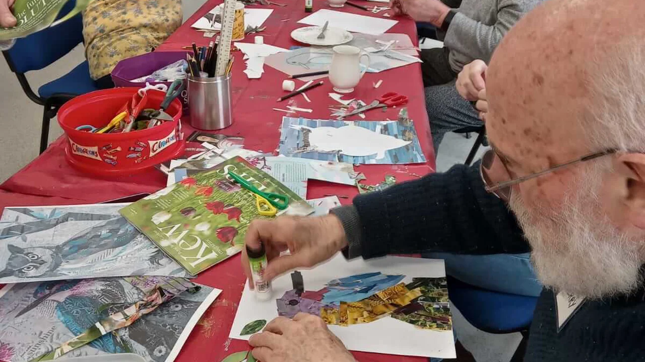 There is a table with a red table cloth covered with papers and magazines. Around the table are men and women are seated all cutting, sticking and making collages, taken at Warminster Art group