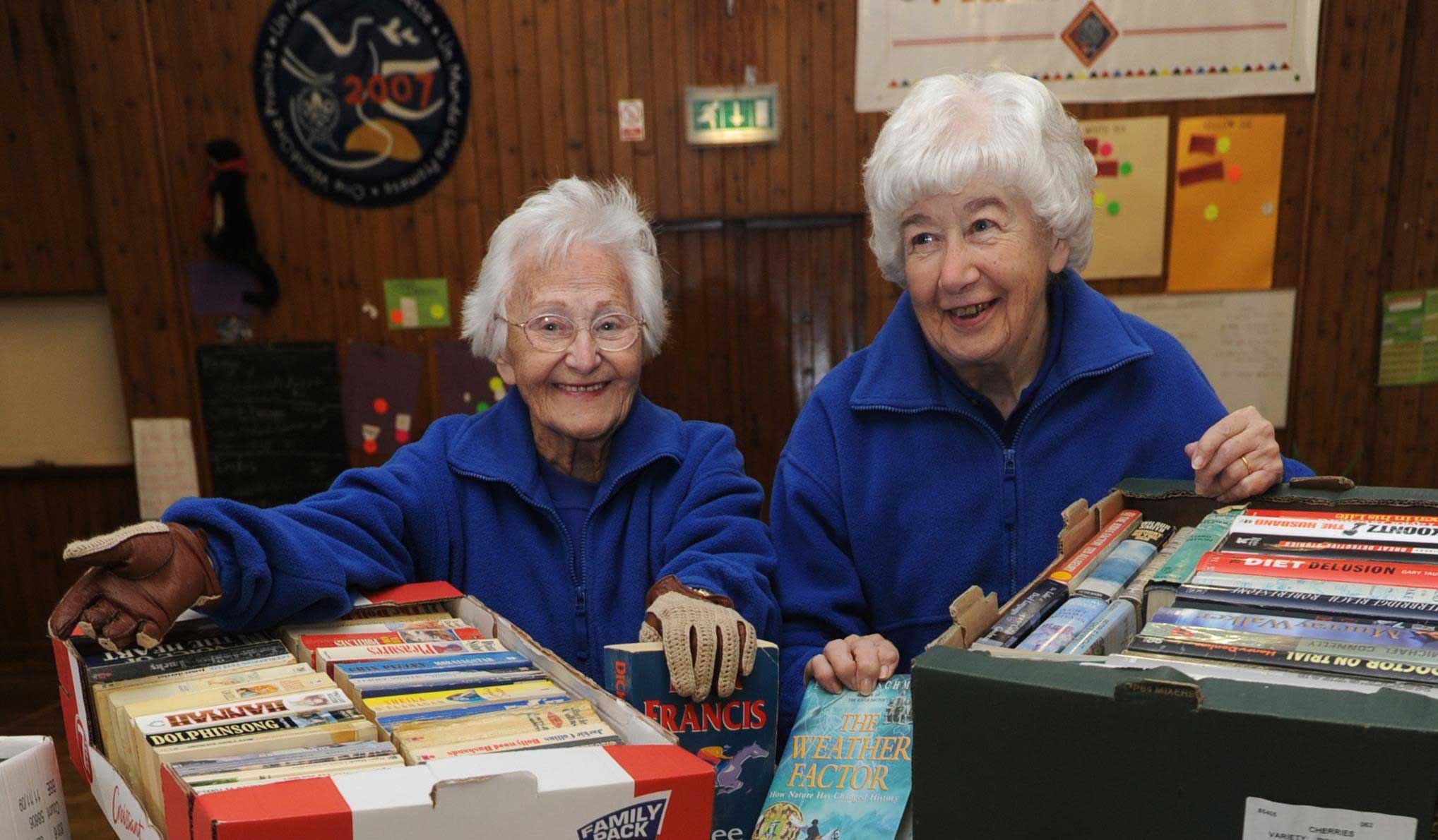 Beryl and a friend in matching blue fleeces with a box of paperback books at a fundraising stall. Both ladies are grey-haired, twinkly-eyed and and smiling