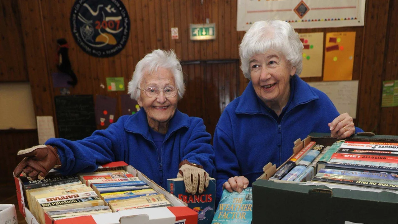 Beryl and a friend in matching blue fleeces with a box of paperback books at a fundraising stall. Both ladies are grey-haired, twinkly-eyed and and smiling