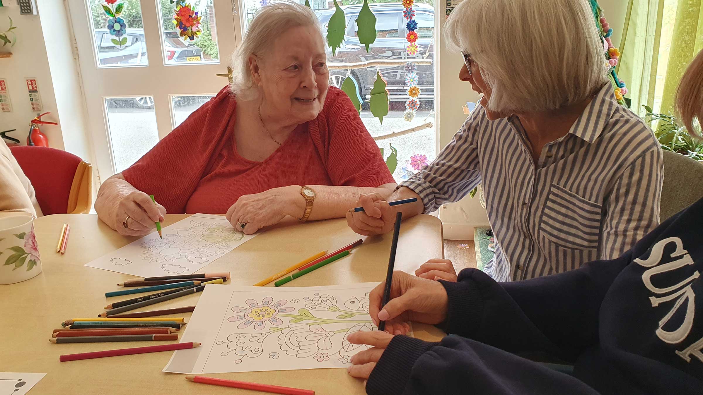 An older lady sitting in an orange top with a younger lady in a stripy shirt, both are colouring in and chatting to each other at the same time, with colouring pencils on the table nearby