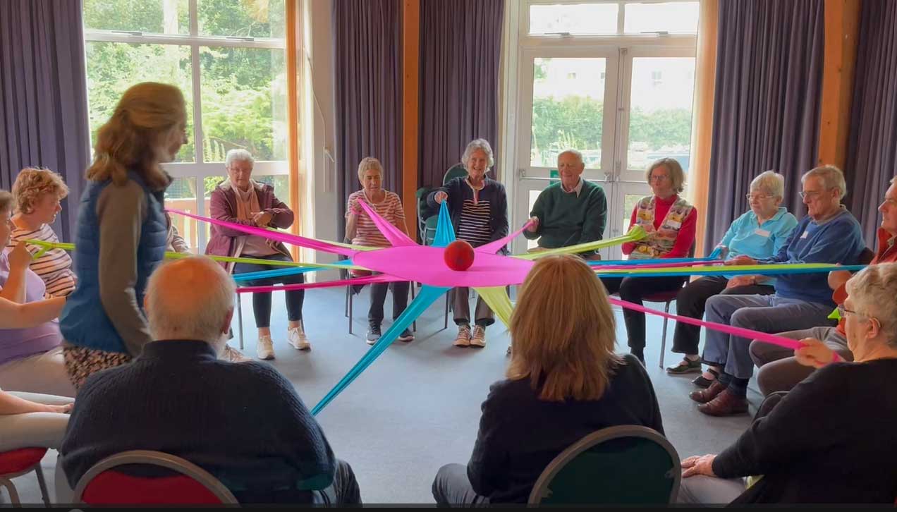 A large group of older people sitting pulling on red, yellow, green and blue stretchy bands joined in the middle and balancing a ball, all smiling at Devizes Movement for the Mind