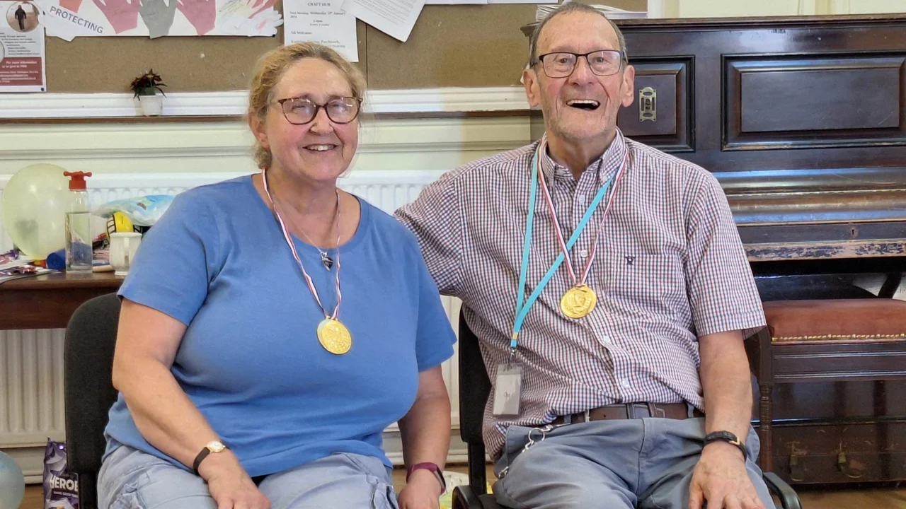 A man and a lady seated both wearing gold medals and glasses, both smiling at the Amesbury Movement group