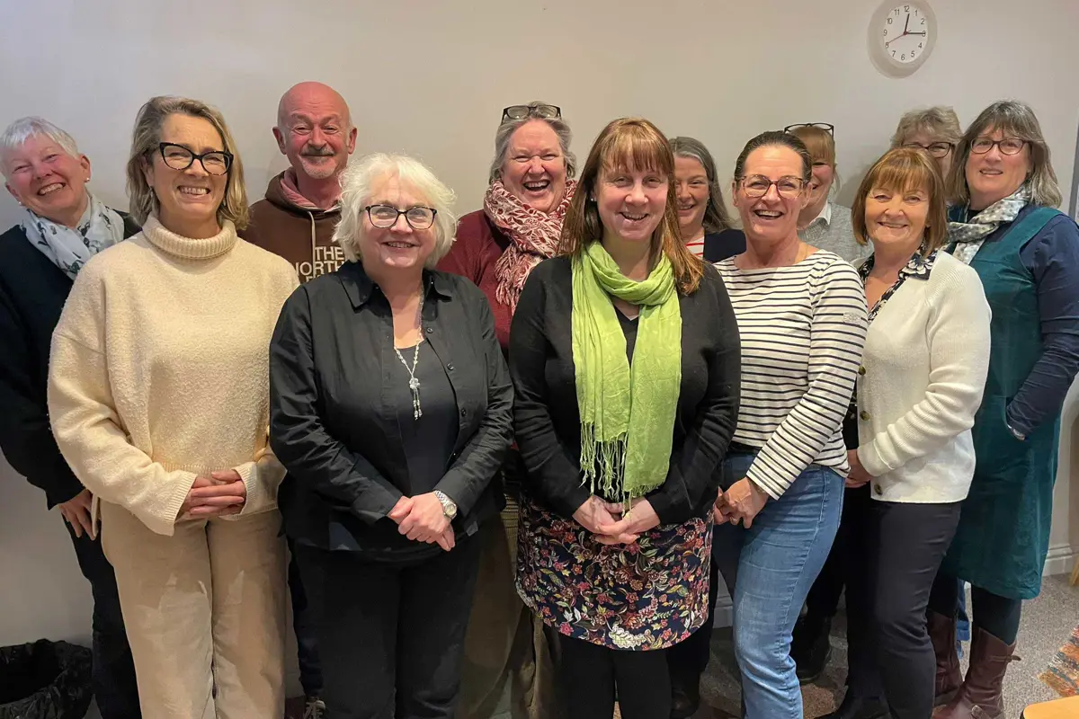 Group of 12 dementia advisers standing together in a meeting room., smiling as they face the camera