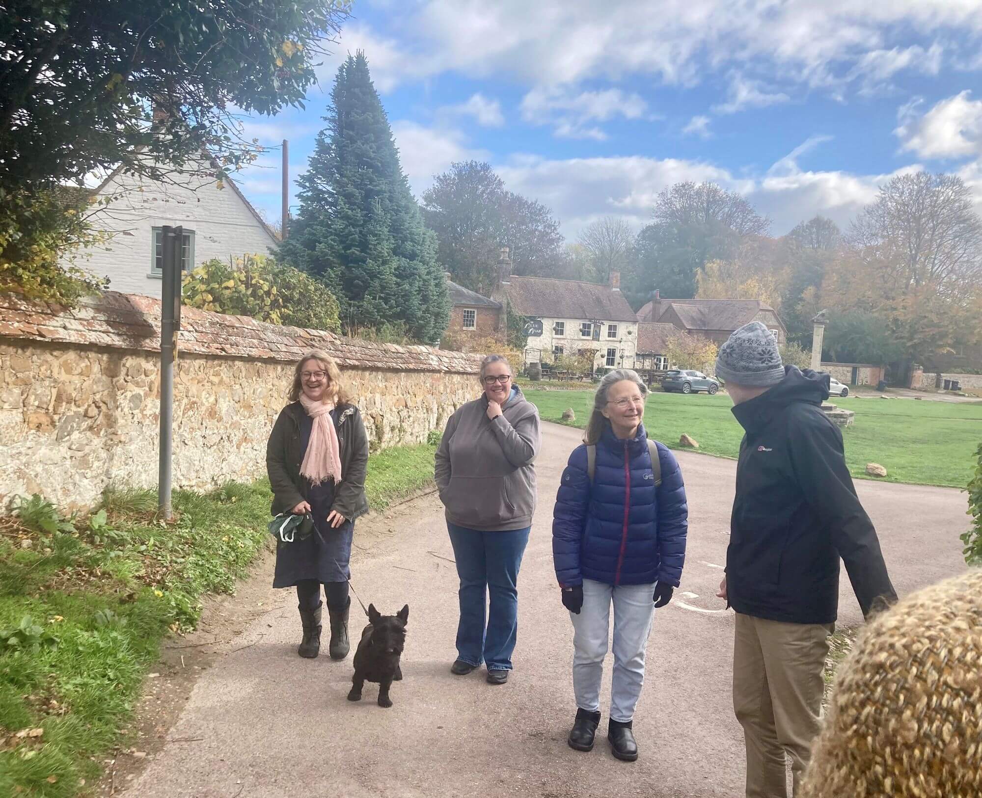 Group of people and a black Scottie dog walking together through a picturesque village. Blue sky with clouds and thatched cottages in the baackground.