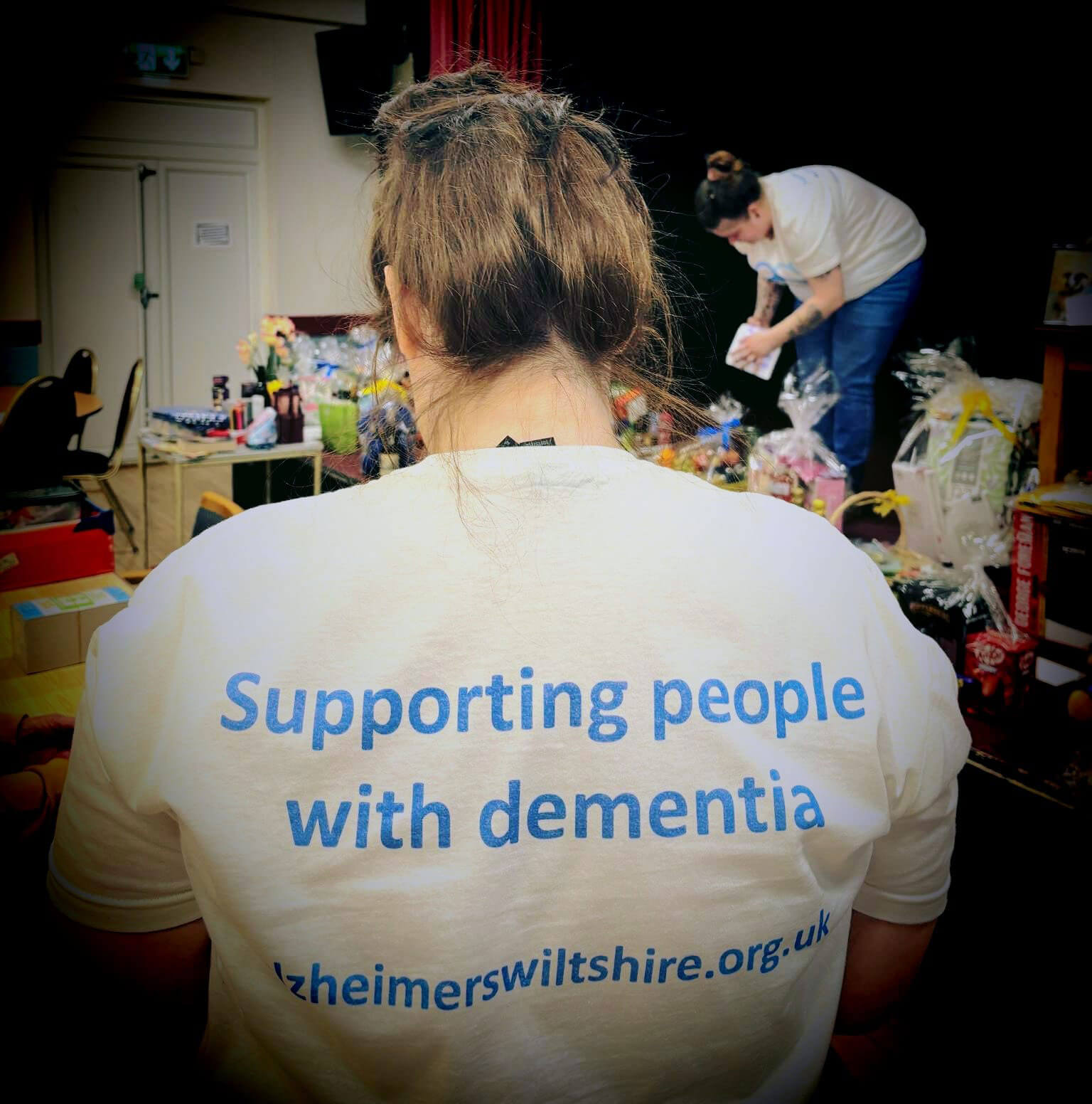 A lady standing with her back to us, wearing a white T shirt which says 'Supporting people with dementia' and Alzheimers Support web site address.  Another lady in the background is leaning over raffle prizes on a table 