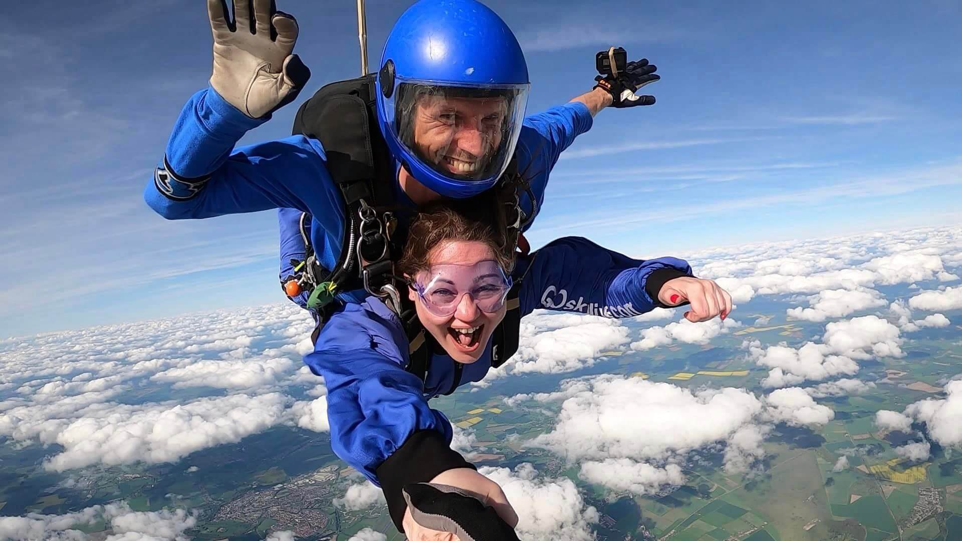 A face on shot of two people in blue suits doing a tandem sky dive both smiling and laughing with blue sky and clouds below them