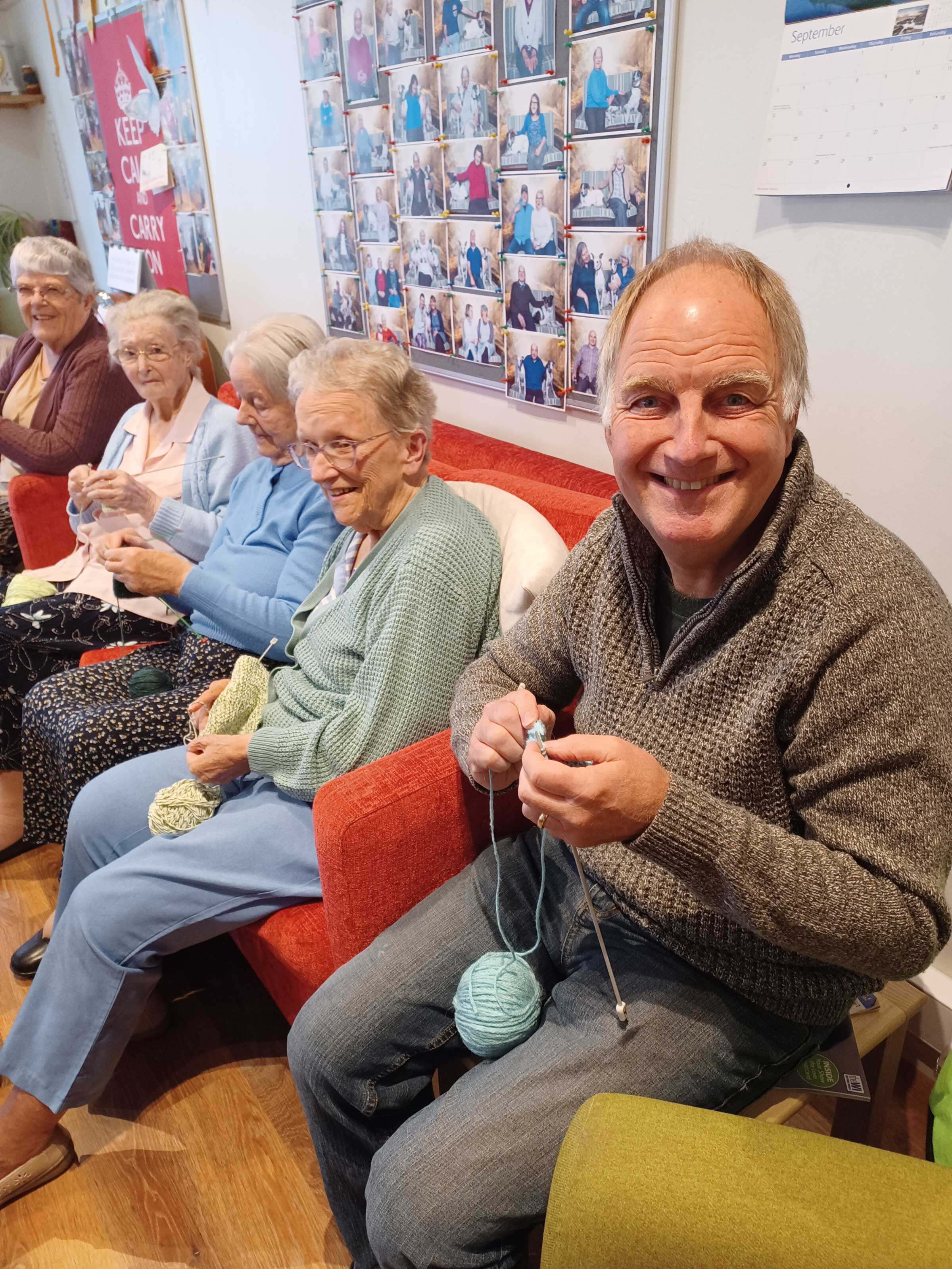 Volunteer Stephen wearing a grey jumper is smiling at us and sitting next to a line of four ladies in arm chairs, all have knitting needles and wool and are concentrating on their knitting 