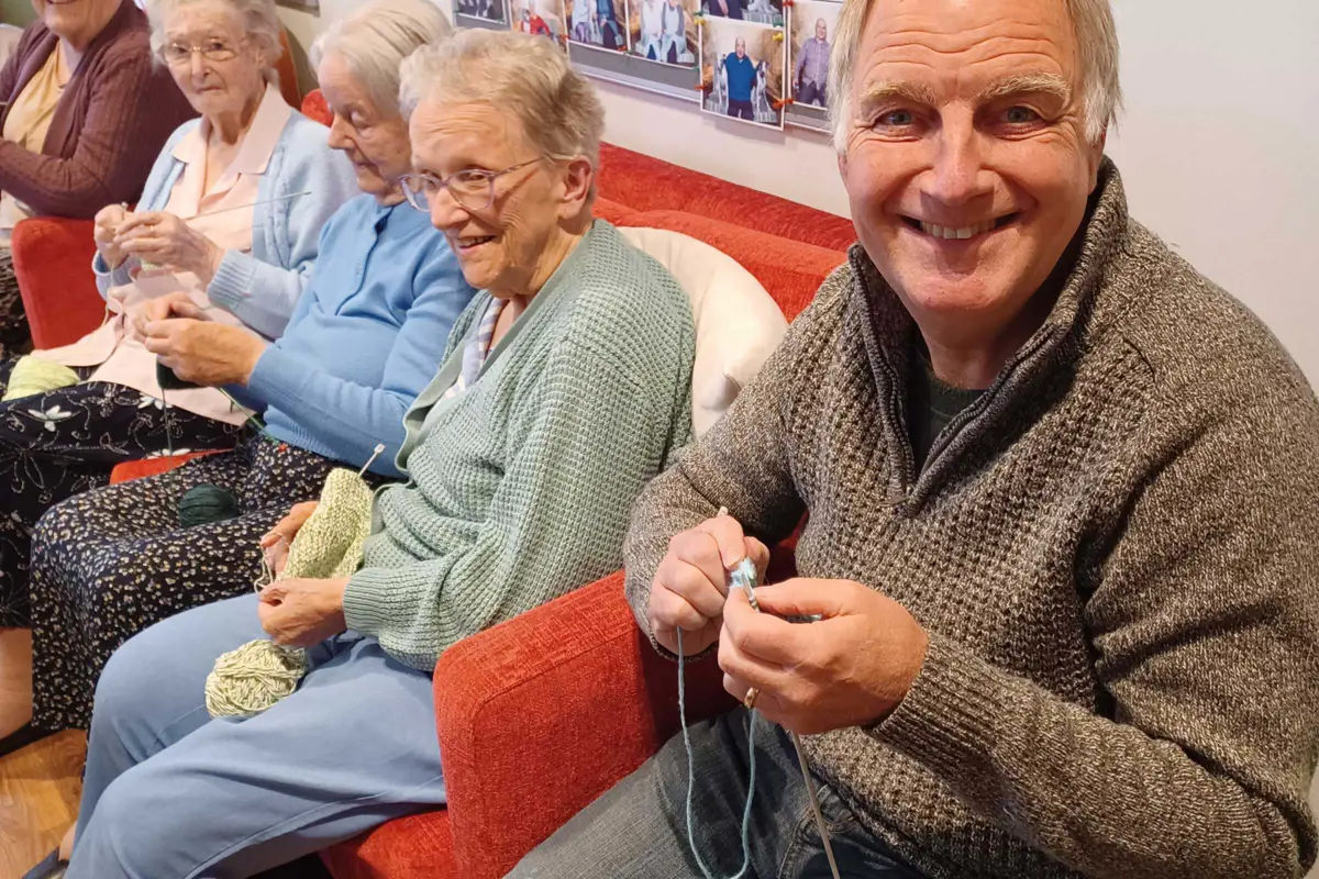 A volunteer, Stephen wearing a grey jumper is smiling at us and sitting next to a line of four ladies in arm chairs, all have knitting needles and wool and are concentrating on their knitting