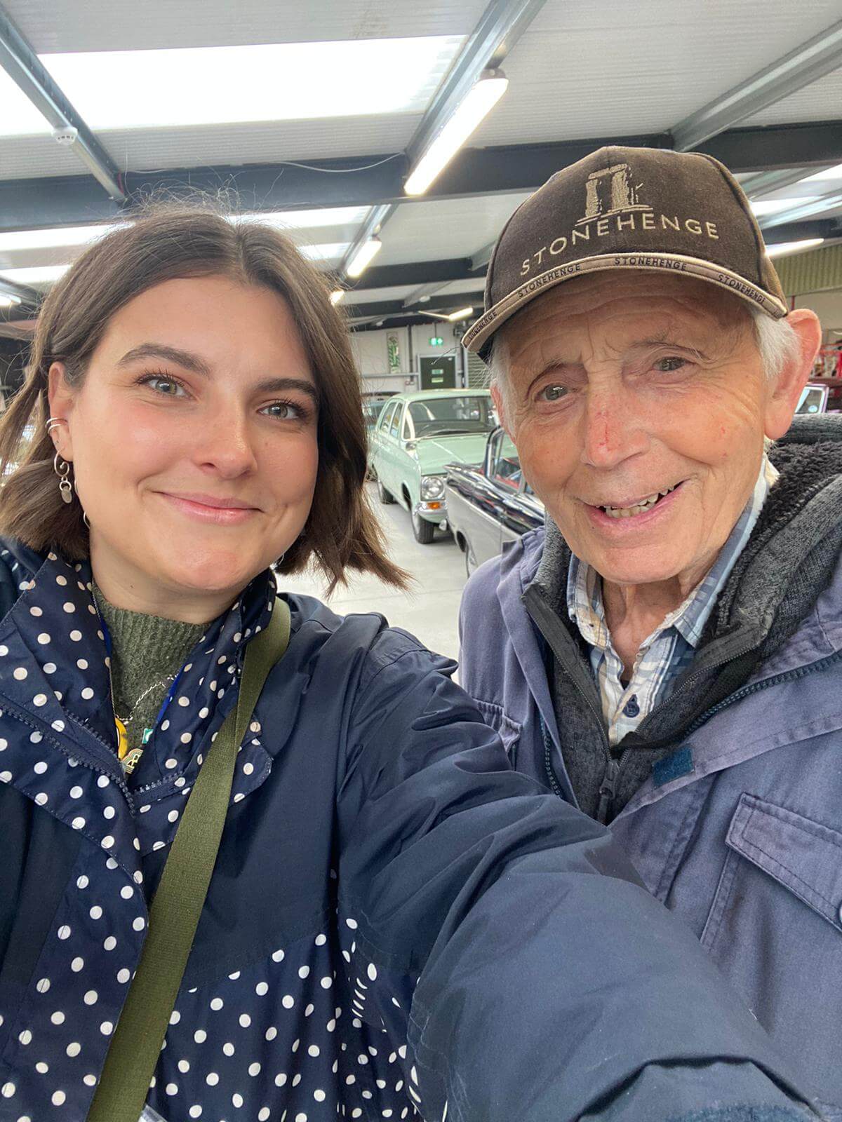 A close up of a younger lady with brown hair and a spotty coat standing with an older man with a Stonehenge baseball cap and a blue coat.  They are in a bit room with classic cars behind them
