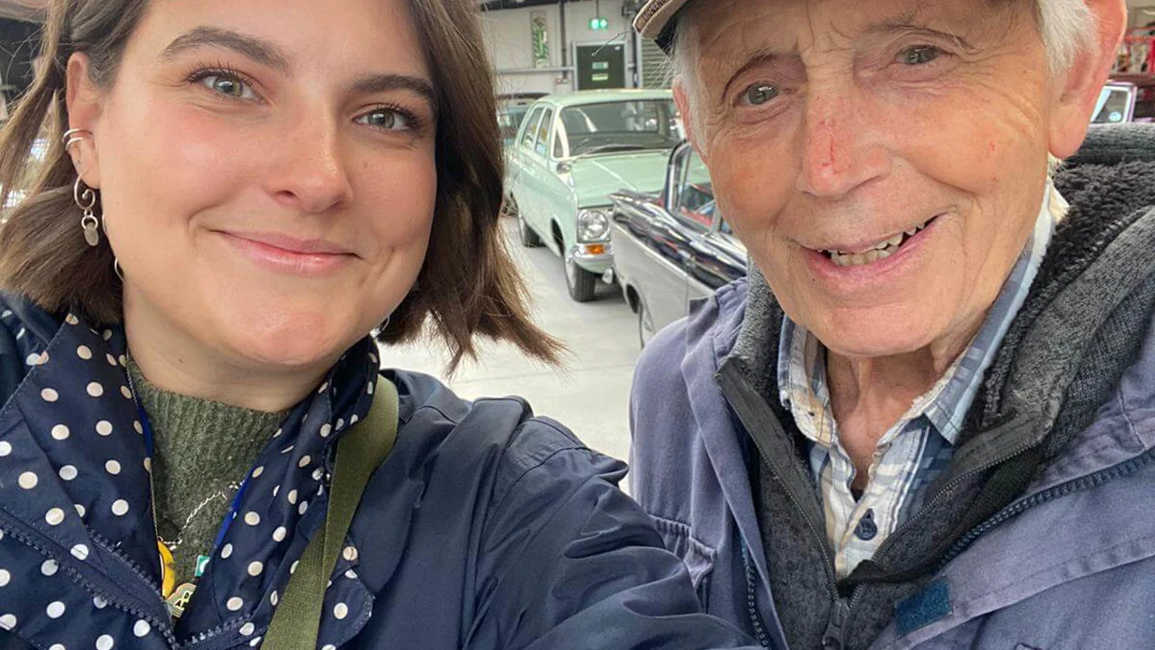 A close up of a younger lady with brown hair and a spotty coat standing with an older man with a Stonehenge baseball cap and a blue coat. They are in a bit room with classic cars behind them