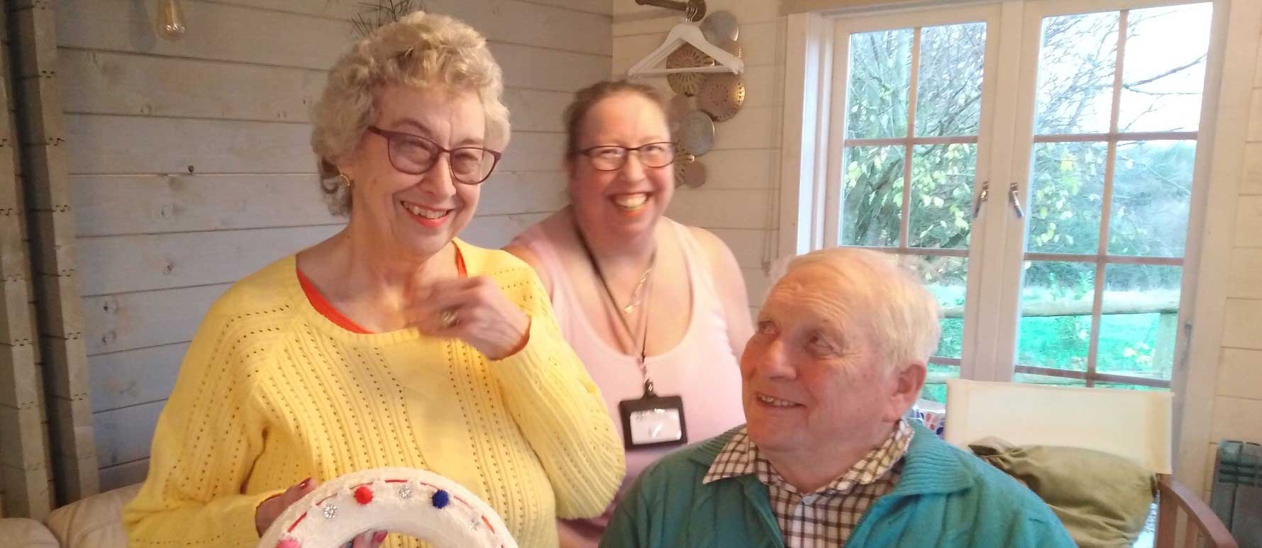 An older lady laughing, wearing a yellow jumper with an older man looking at her and smiling with a younger lady behind them wearing a white vest top and also laughing 