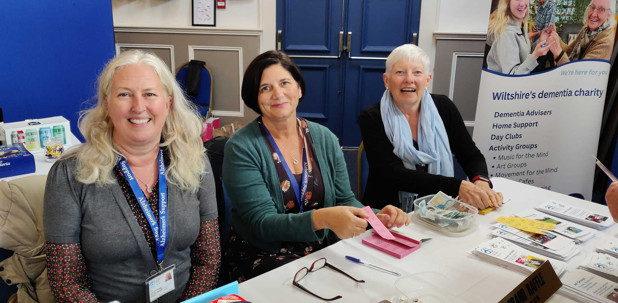 Kate flanked by colleagues Sarah and Tina, smiling as they sell raffle tickets behind a table at a fundraising event