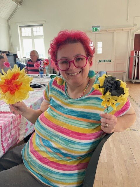 A lady with pink highlights in her hair is sitting smiling at the camera holding two yellow tissue paper flowers on sticks.  She is wearing a pink, yellow and blue striped T shirt.  Taken at Chippenham Art group 