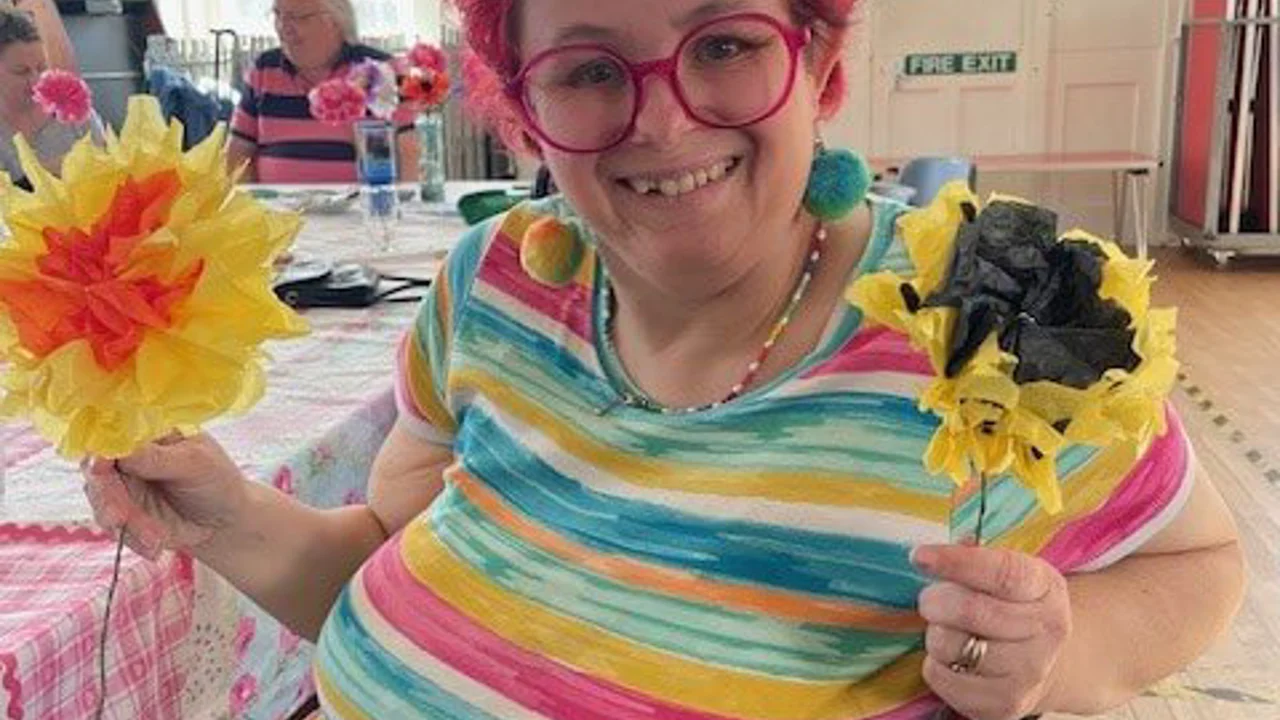 A lady with pink highlights in her hair is sitting smiling at the camera holding two yellow tissue paper flowers on sticks. She is wearing a pink, yellow and blue striped T shirt. Taken at Chippenham Art group