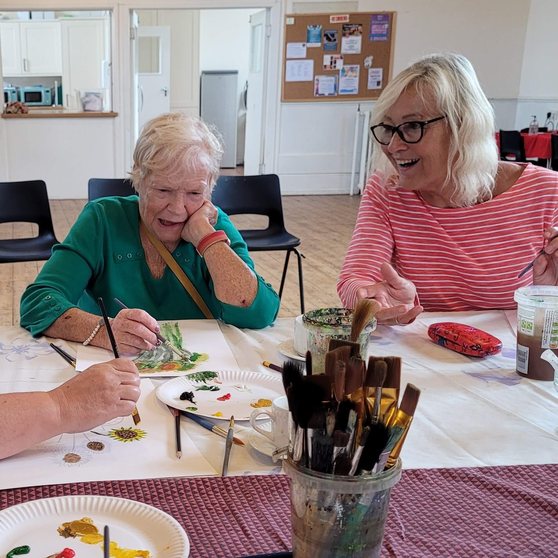 A very happy smiling lady wearing a red and white striped shirt is sitting at an art table.  There is a lady next to her painting and concentrating on her art and in the foreground there is plates with paint and paint brushes