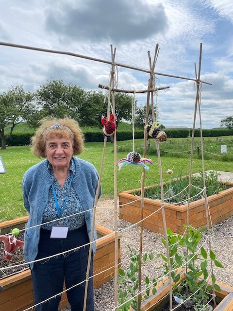A lady in a blue cardigan is standing next to a wooden frame with sweet peas growing up it.  There are crocheted insects hanging off the frame just above the ladies head and raised beds in the background. Taken at REME Muddy Boots group 