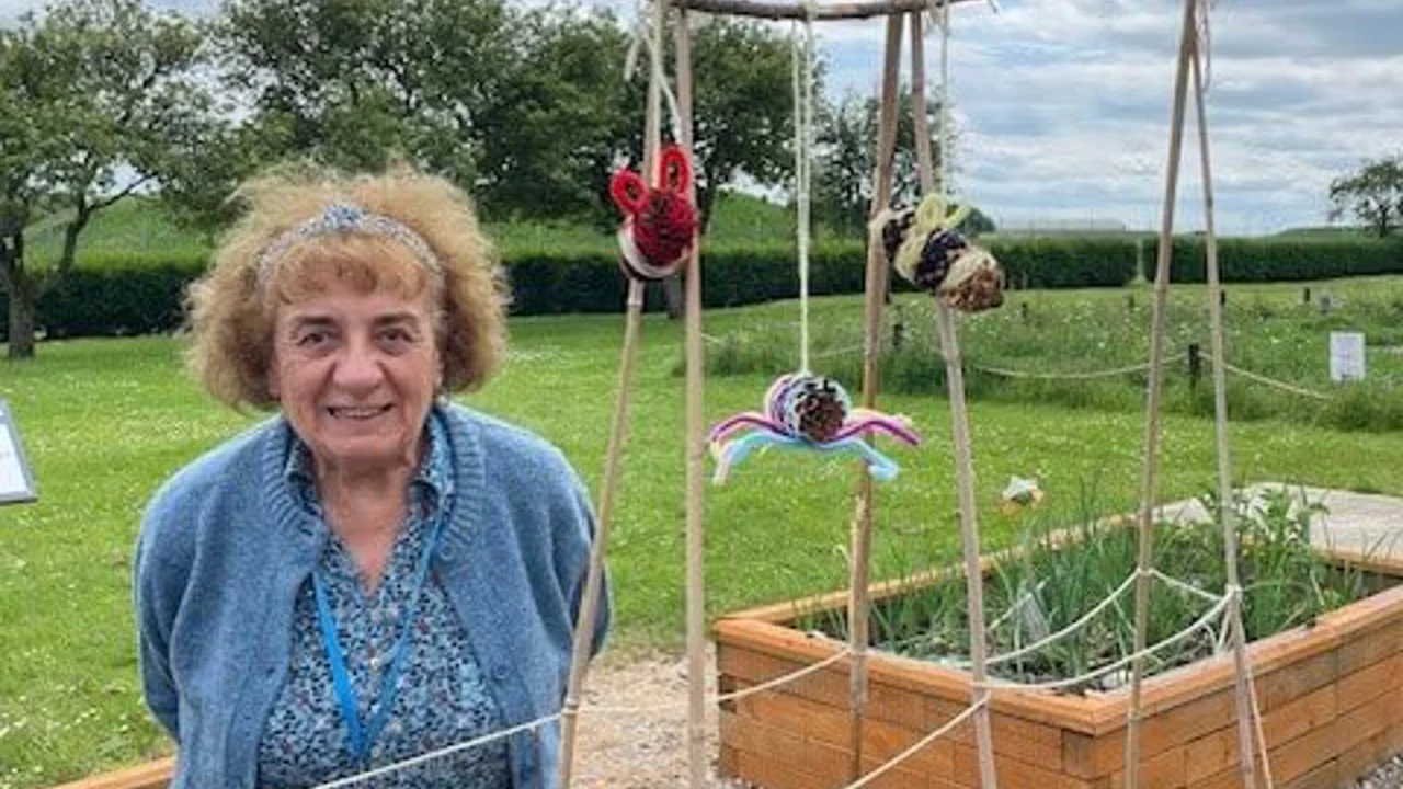 A lady in a blue cardigan is standing next to a wooden frame with sweet peas growing up it. There are crocheted insects hanging off the frame just above the ladies head and raised beds in the background. Taken at REME Muddy Boots group