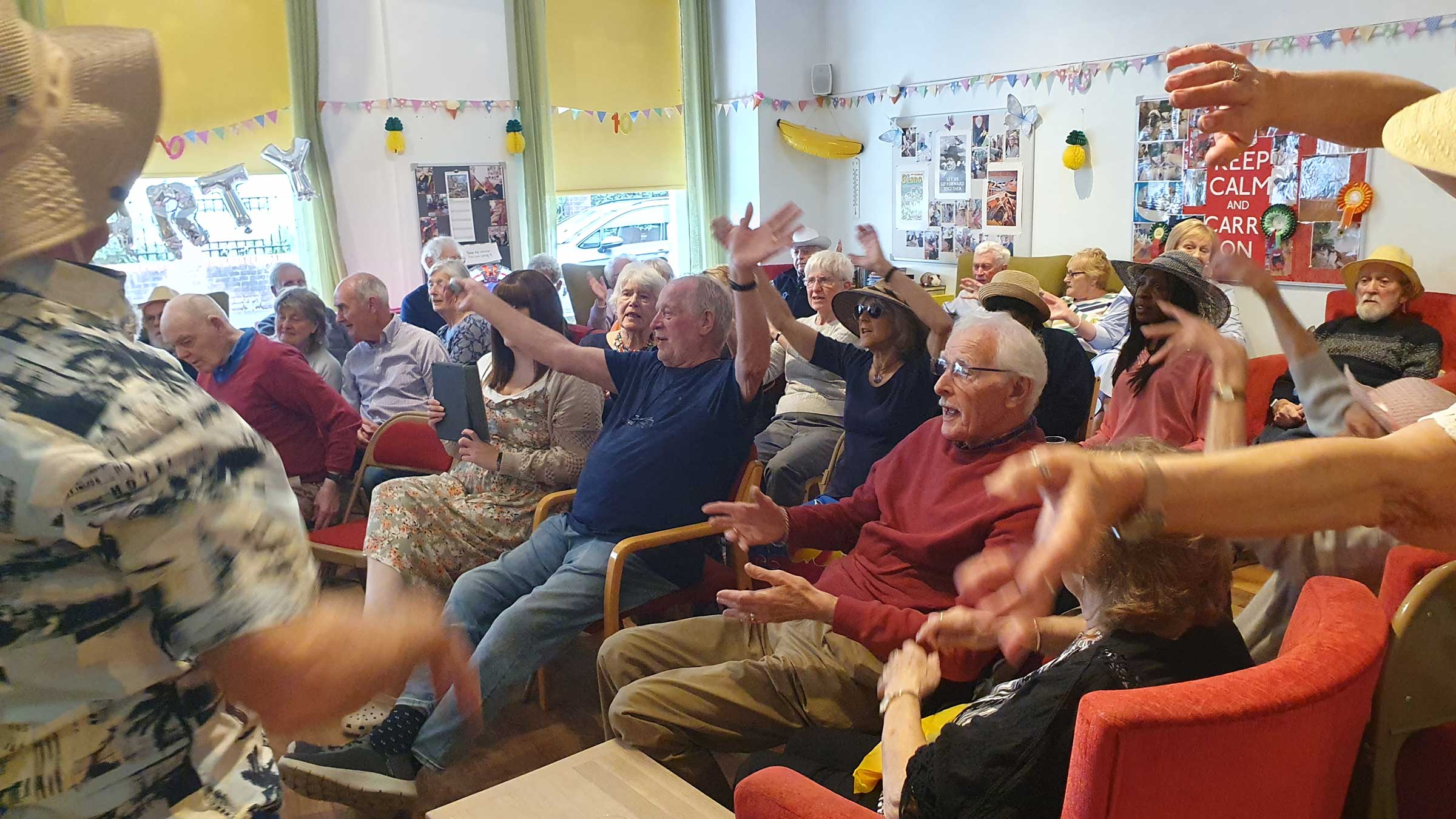 About 20 people sitting, clapping and waving their arms as they listen to music at the party.