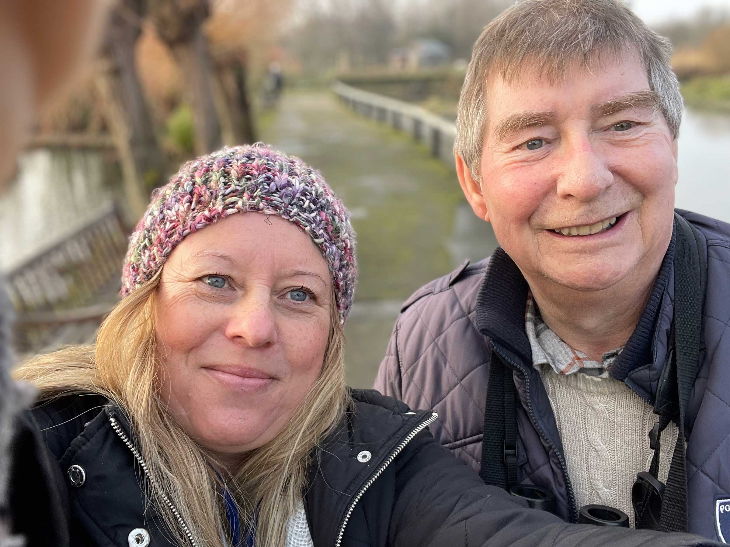 This is a close up of a lady in a pinky grey beanie hat with blond hair and a man in a grey jacket standing together on a path next to some water