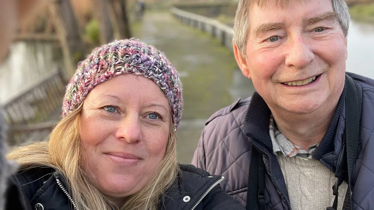 This is a close up of a lady in a pinky grey beanie hat with blond hair and a man in a grey jacket standing together on a path next to some water