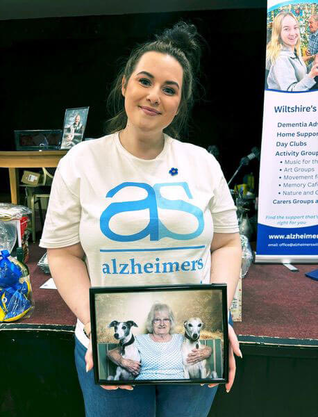 A lady smiling with dark hair wearing a white Alzheimer's Support T shirt holding a picture of a lady wearing a spotty top with her arms around two whippets.