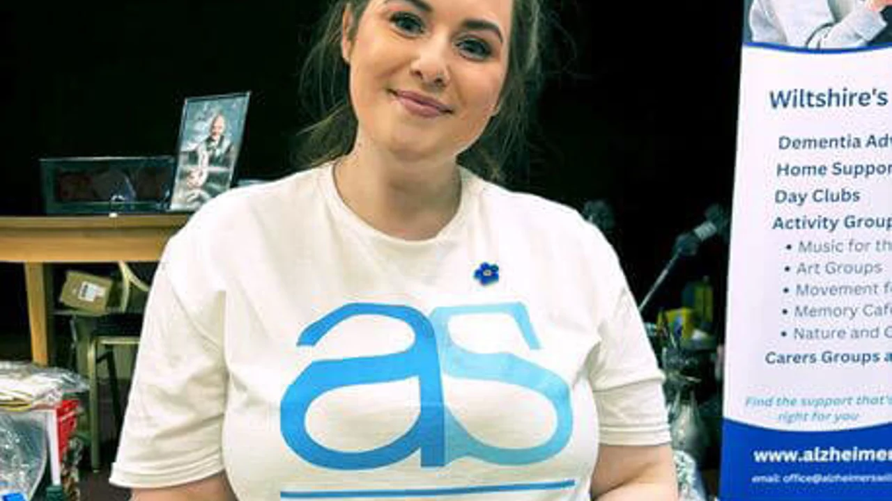 A lady smiling with dark hair wearing a white Alzheimer's Support T shirt holding a picture of a lady wearing a spotty top with her arms around two whippets.