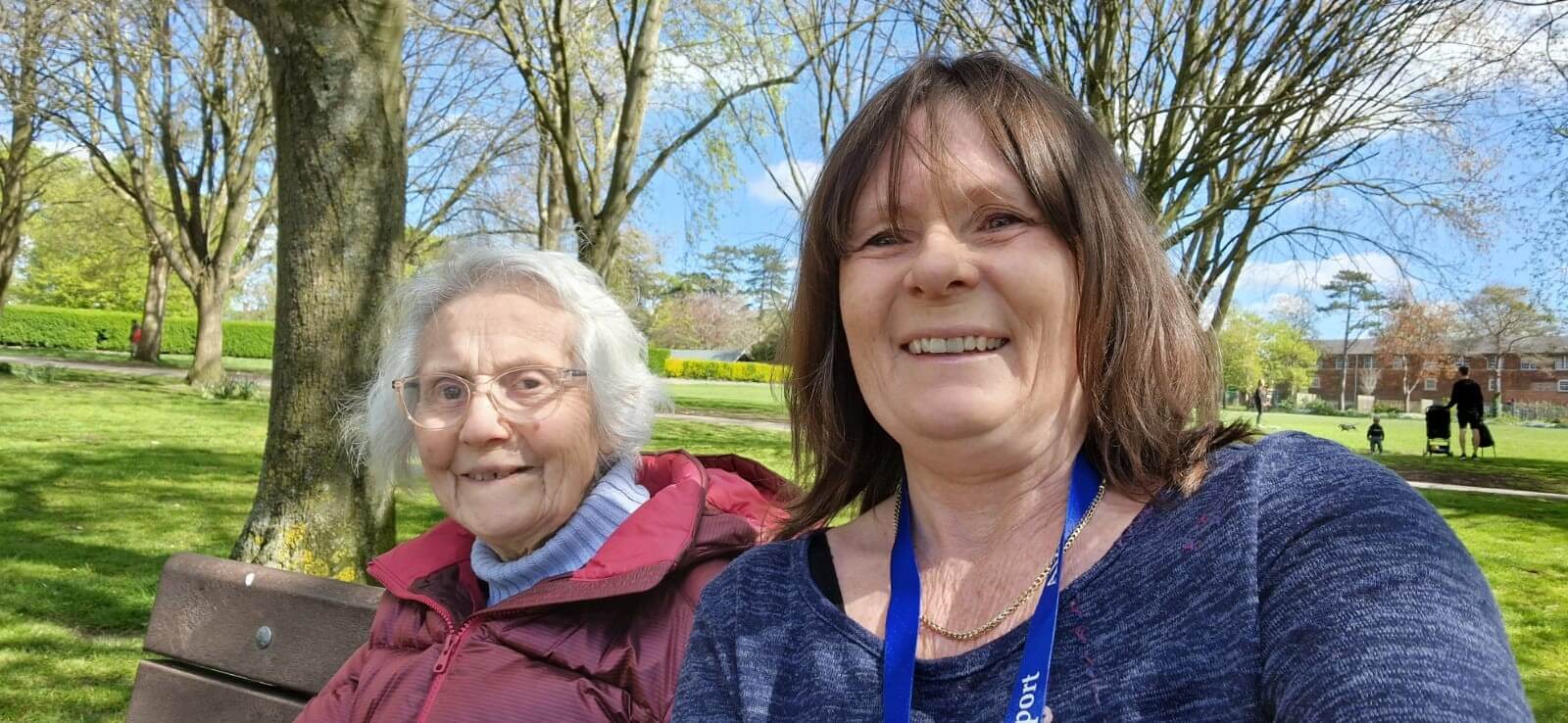 A younger lady in a blue top with brown hair and a lady in a pink coat and grey hair sit on a bench in a park with trees and grass.  