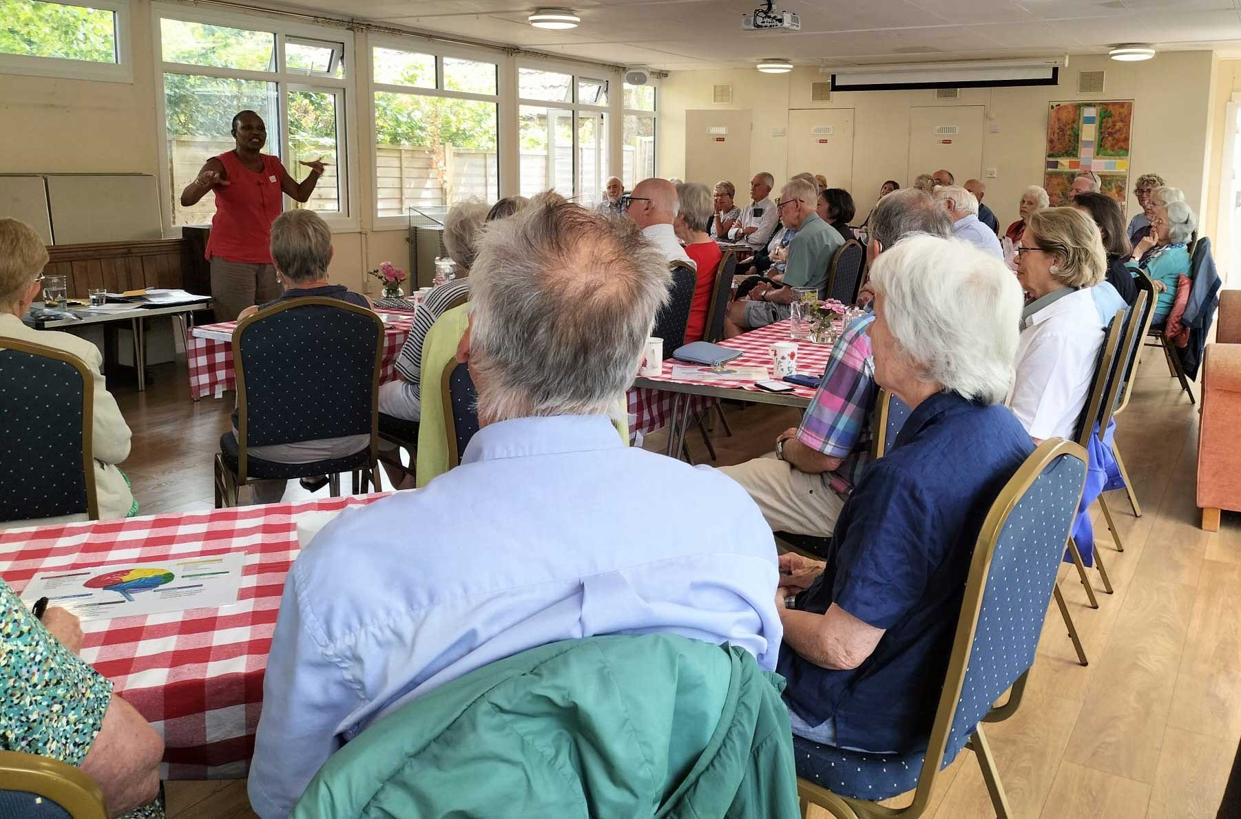 A large room at Winsley Church is shown full of many people all sat facing to the front of the room, where a lady is standing presenting a talk.  There are windows and a table behind her 