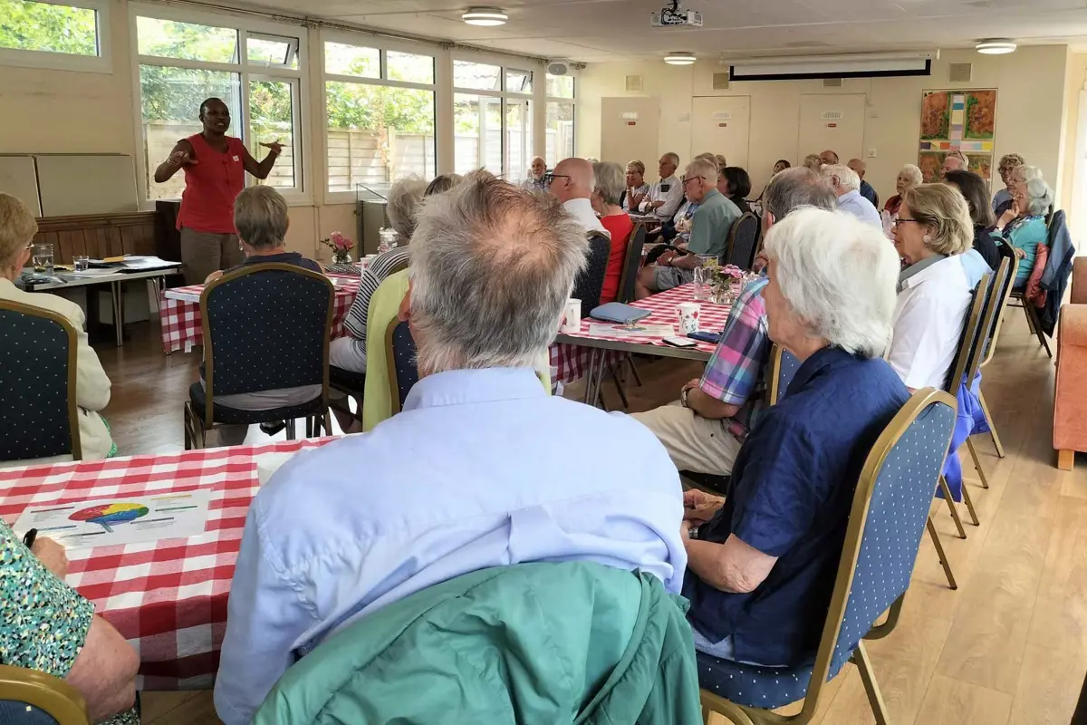 A large room at Winsley Church is shown full of many people all sat facing to the front of the room, where a lady is standing presenting a talk.  There are windows and a table behind her 