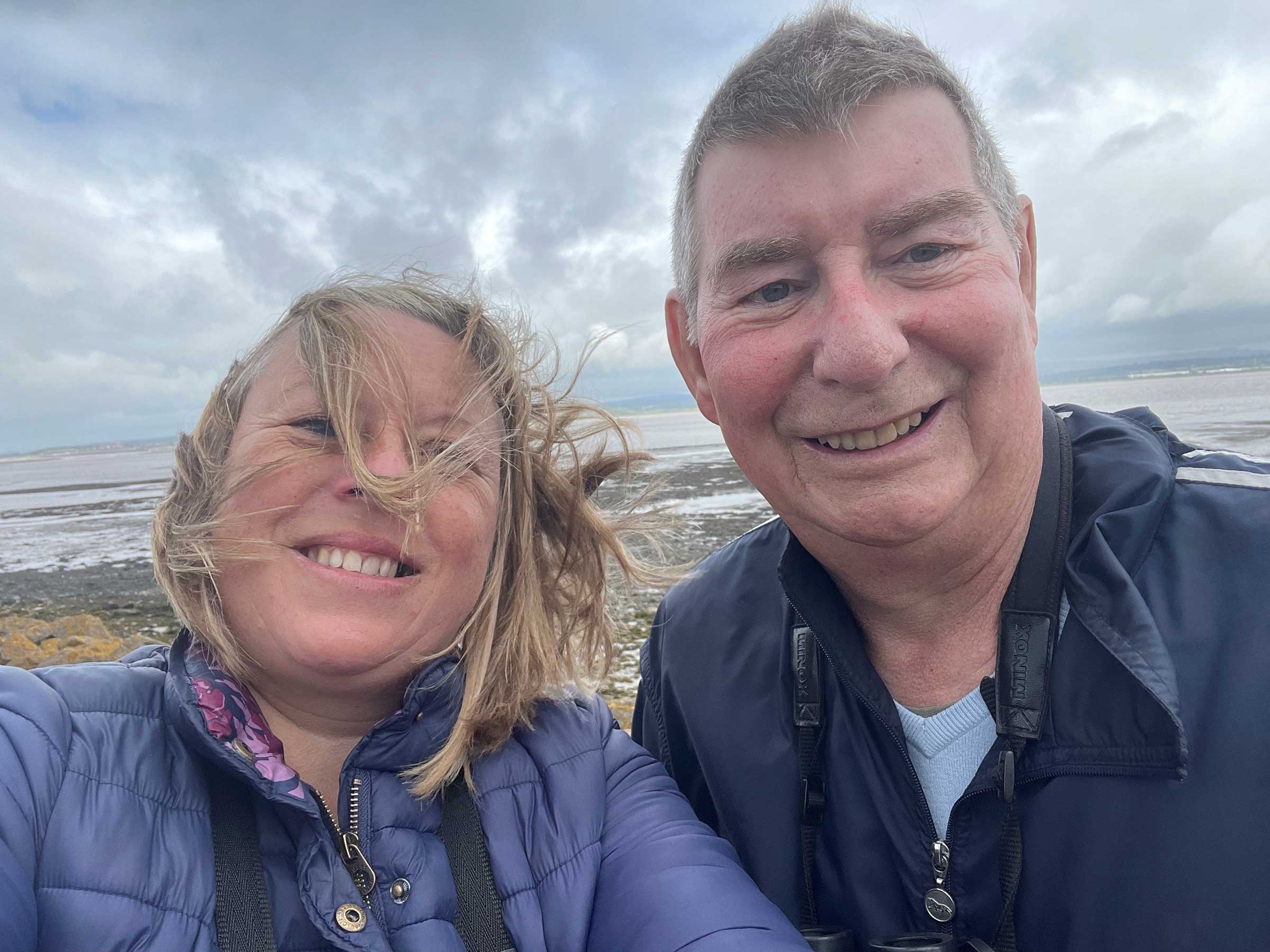 This is a close up, selfie, with an older man and a younger woman.  The wind is blowing the ladies hair over her face and there is an open seascape behind them