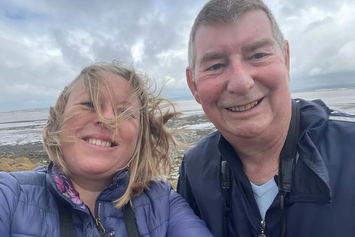 This is a close up, selfie, with an older man and a younger woman. The wind is blowing the ladies hair over her face and there is an open seascape behind them