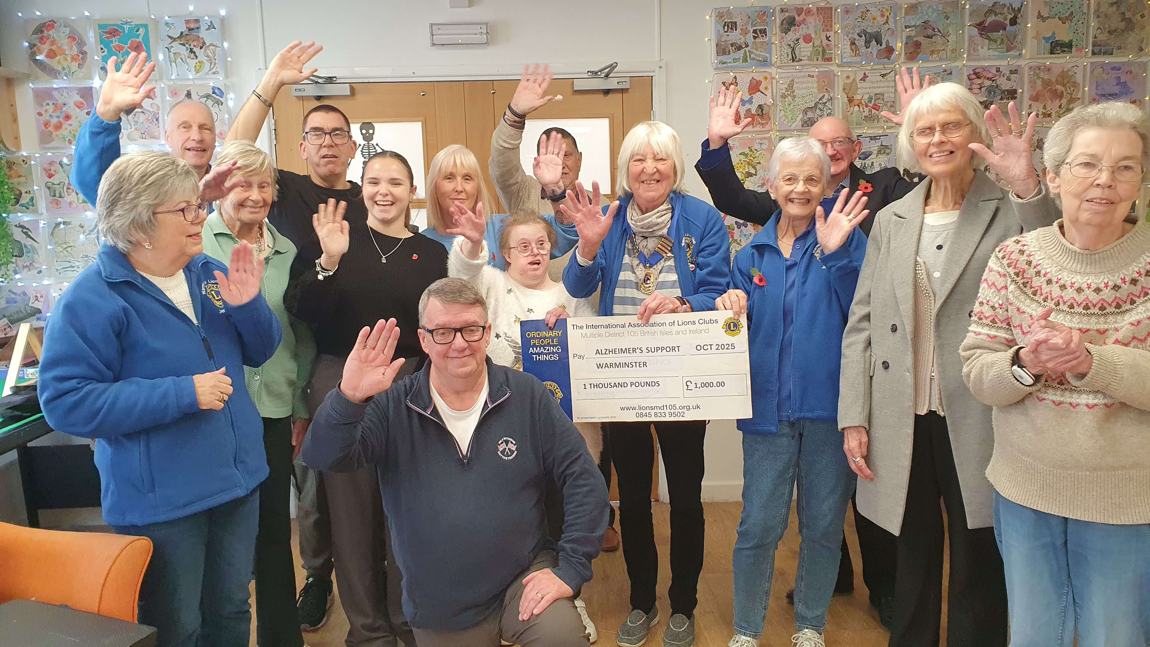 Group of Old Silk Work members and Lions members waving at the camera and holding a large cheque made out for £1,000
