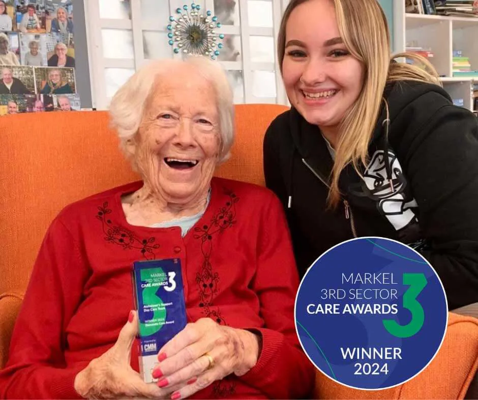 Marjorie and Alex sitting together in Old Silk Works holding the Markel Care Sector award Winner 2024 trophy and smiling