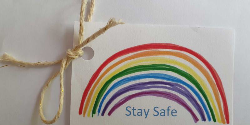 Luggage label with a hand-drawn rainbow, and the words Stay Safe, tied with string in the corner