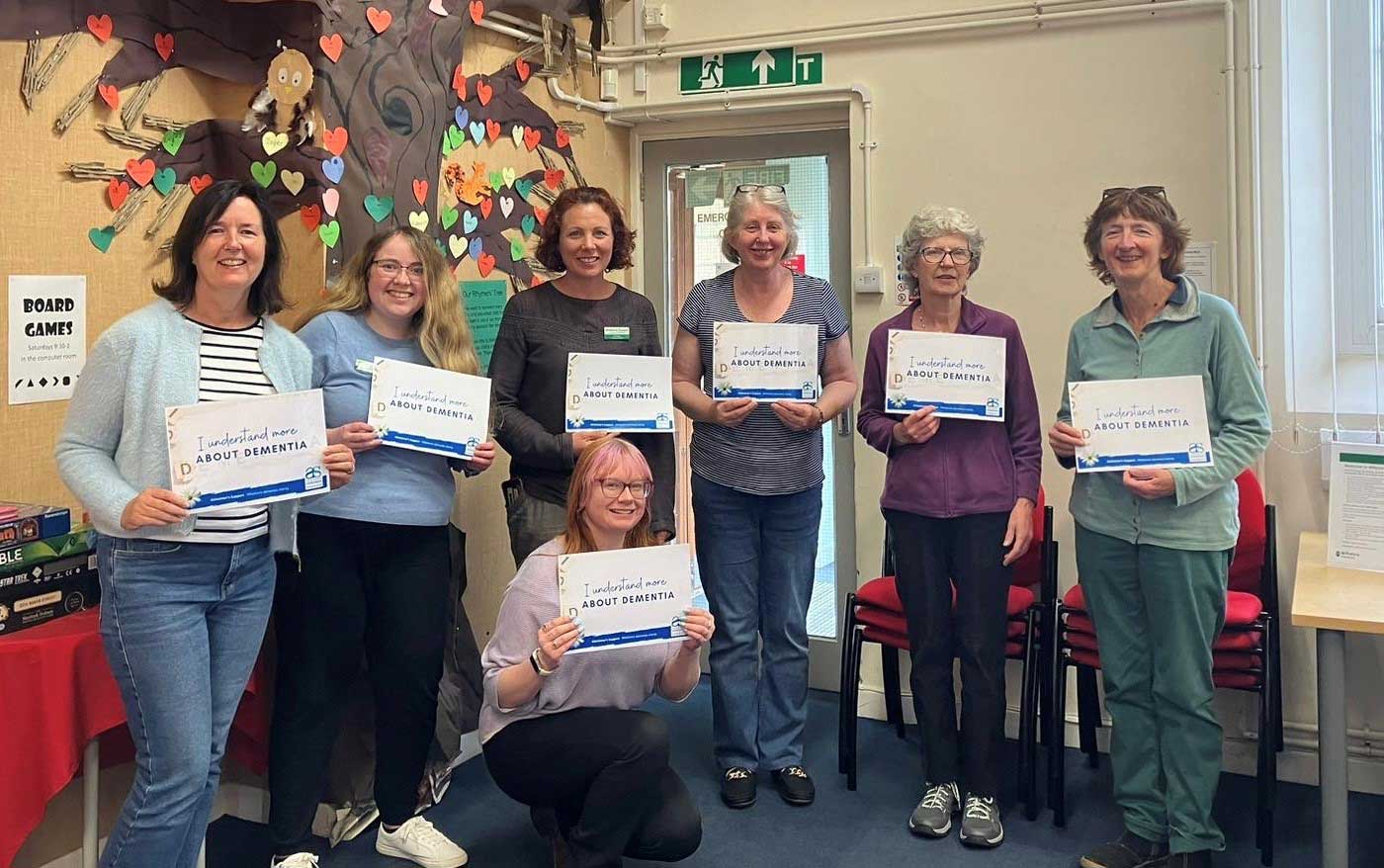A group of people stand together in a semi circle at Malmesbury Library, they all hold a certificate saying I understand more about dementia.  There is a cut out tree with hearts on it behind them
