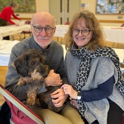 Paul and Laura Willis, sitting facing the camera smiling with their dog Frankie on Laura's lap.