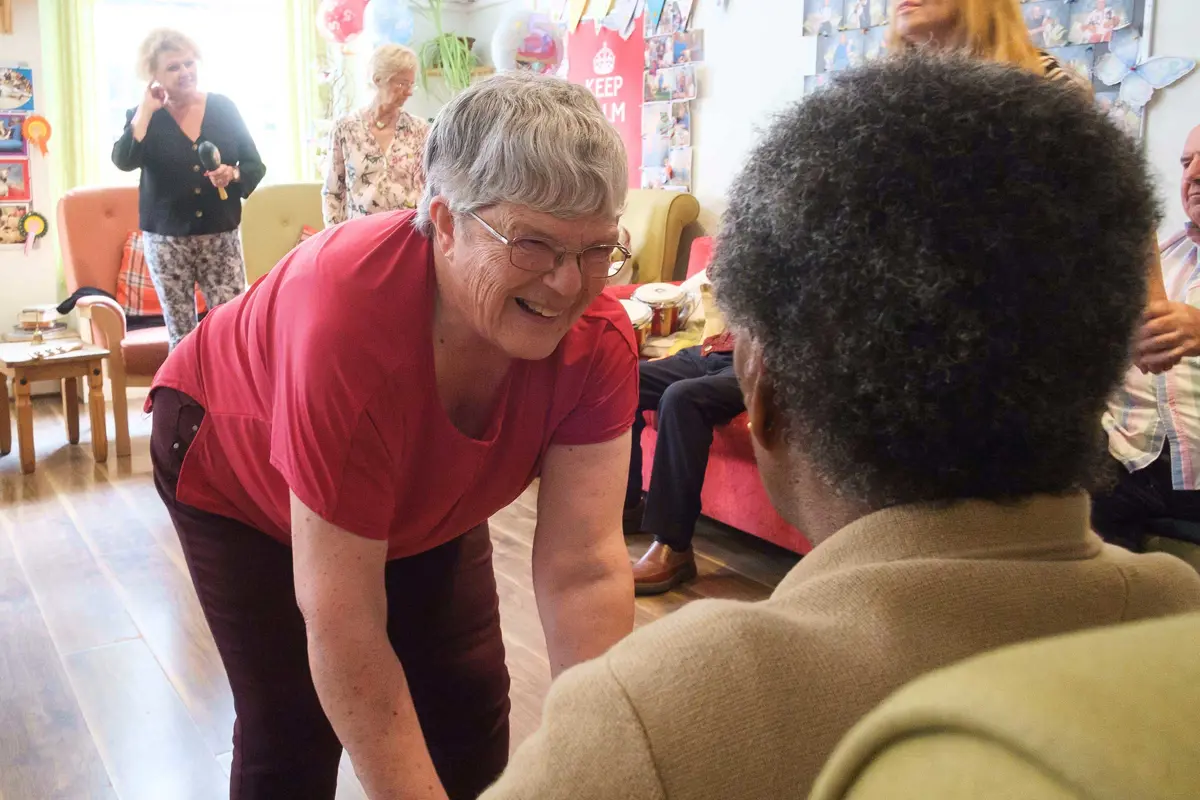 A lady volunteer in red shirt bends forward and smiles to a lady seated in a chair who we see from behind