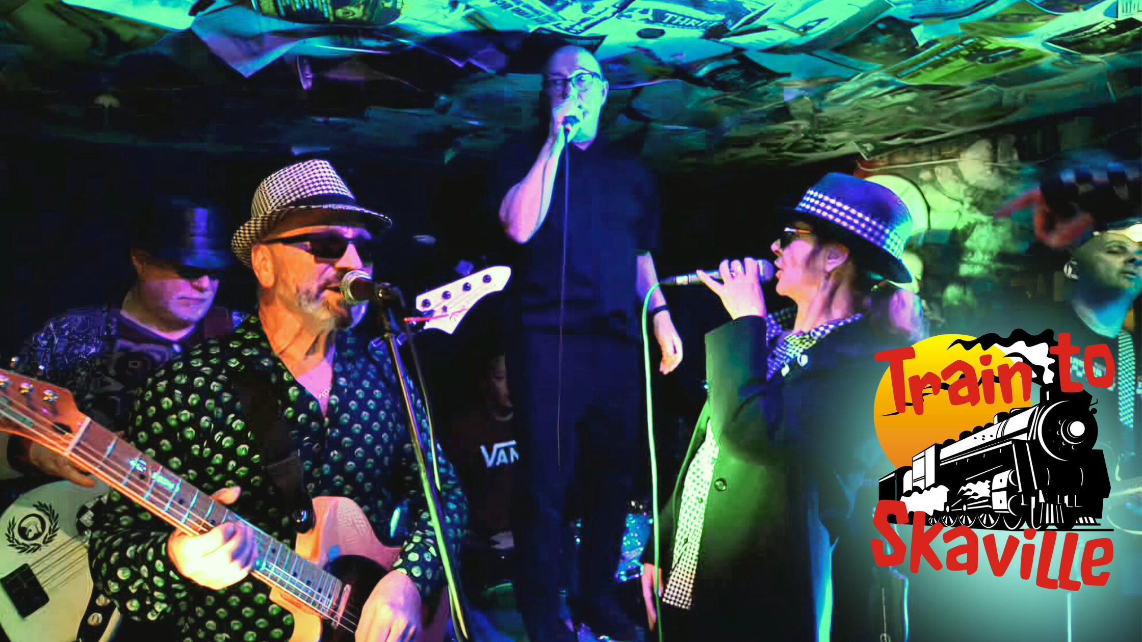 Members of the ska tribute band Train to Skaville performing on stage under neon green and purple spotlights. Their logo containing their name in red with a black and white train in front of an orange sun is displayed on the lower right of the image.