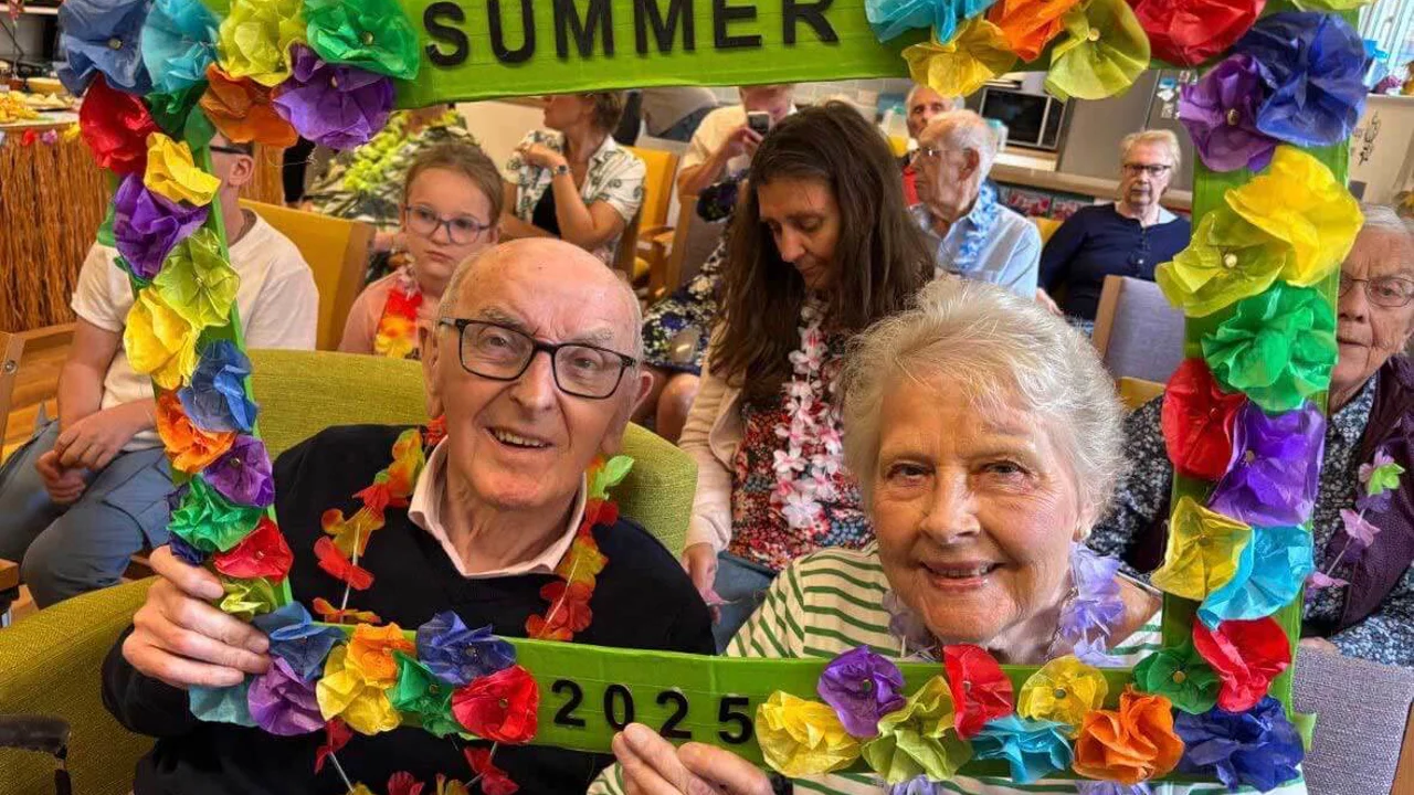 A man and a lady smiling through a frame which says Summer 2025 and is decorated with colourful red, blue, yellow and green flowers