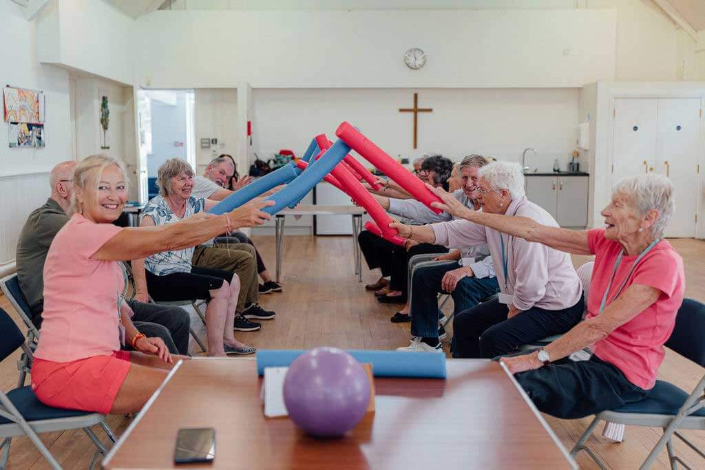 Group of men and women seated opposite each other and holding red and blue foam tubes and making an archway between them at Chippenham Movement for the Mind 