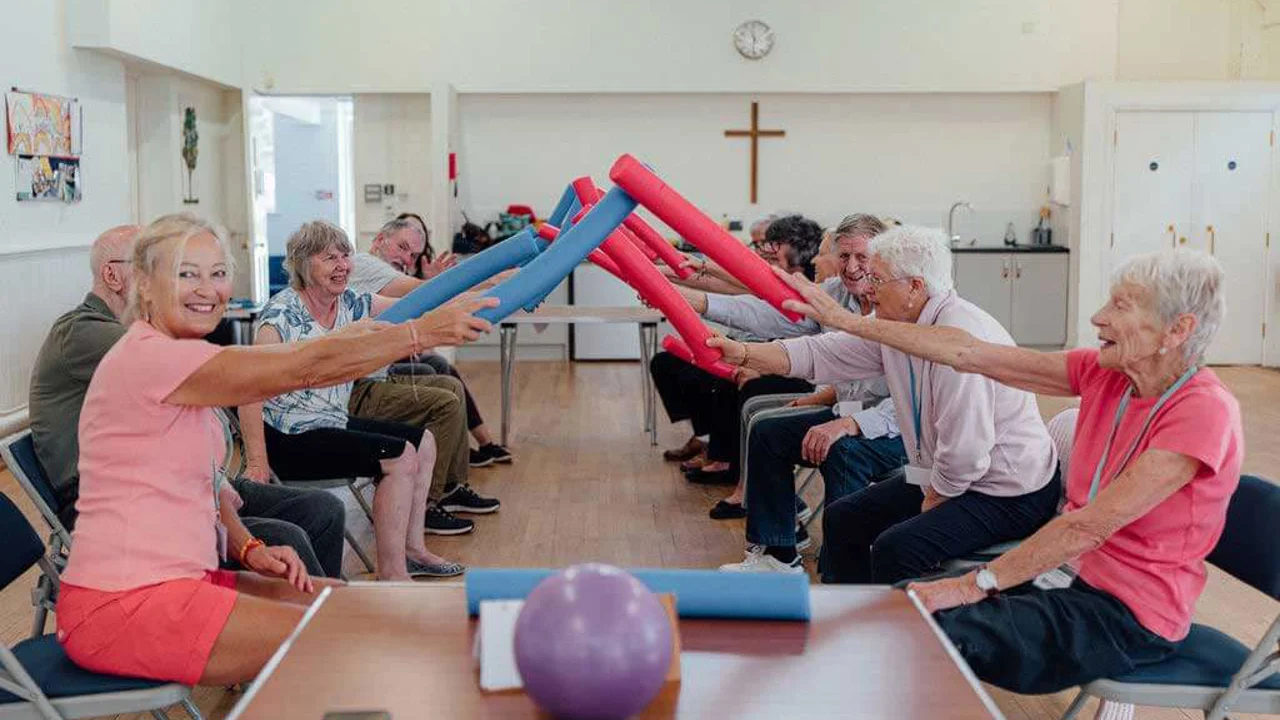 Group of men and women seated opposite each other and holding red and blue foam tubes and making an archway between them at Chippenham Movement for the Mind