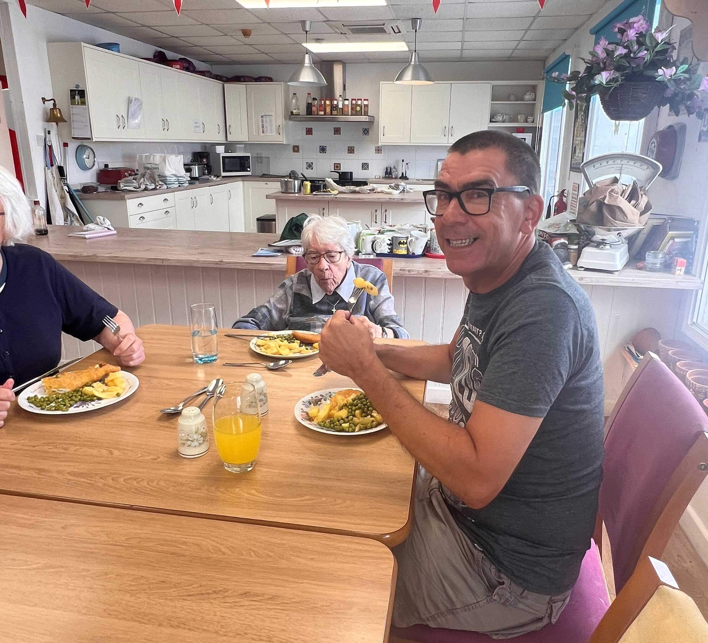 Support worker Tim smiling as he eats a fish and chip lunch at a small table with two club members. The open plan club kitchen is in the background