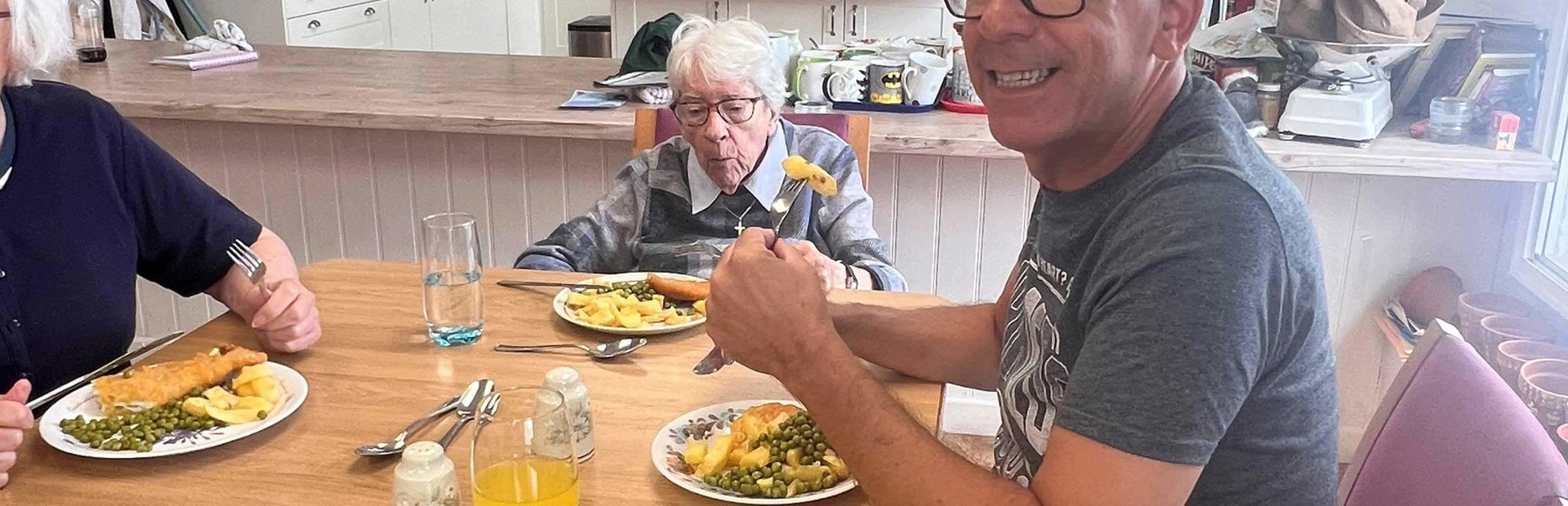 Support worker Tim smiling as he eats a fish and chip lunch at a small table with two club members. The open plan club kitchen is in the background Support worker Tim smiling as he eats a fish and chip lunch at a small table with two club members. The open plan club kitchen is in the background