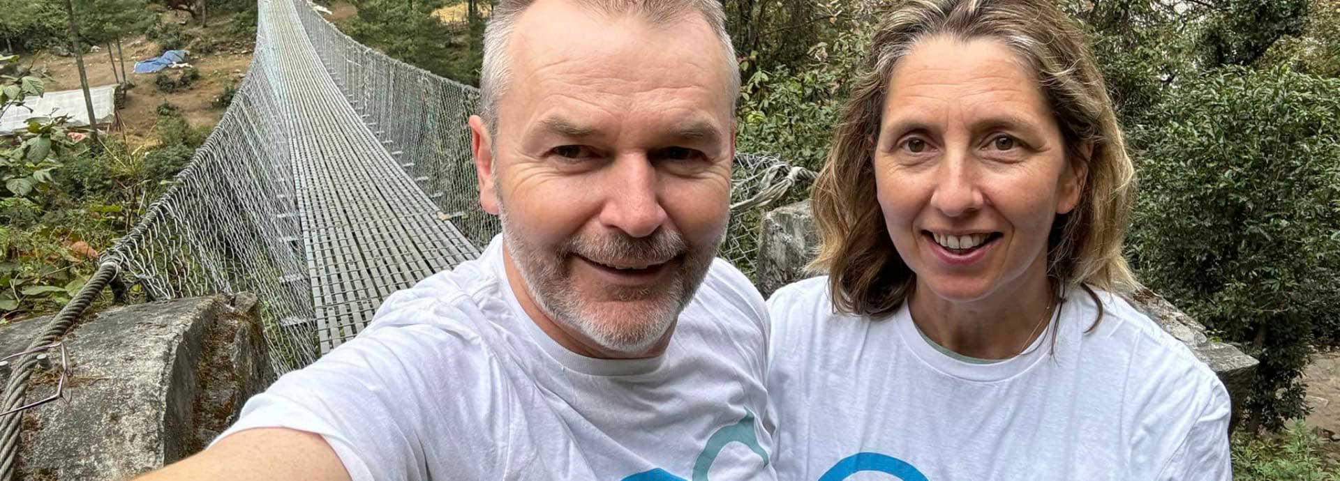A close up selfie taken by Kate and Nigel with a thin rope and wire bridge behind them whilst doing an Everest Base Camp walk