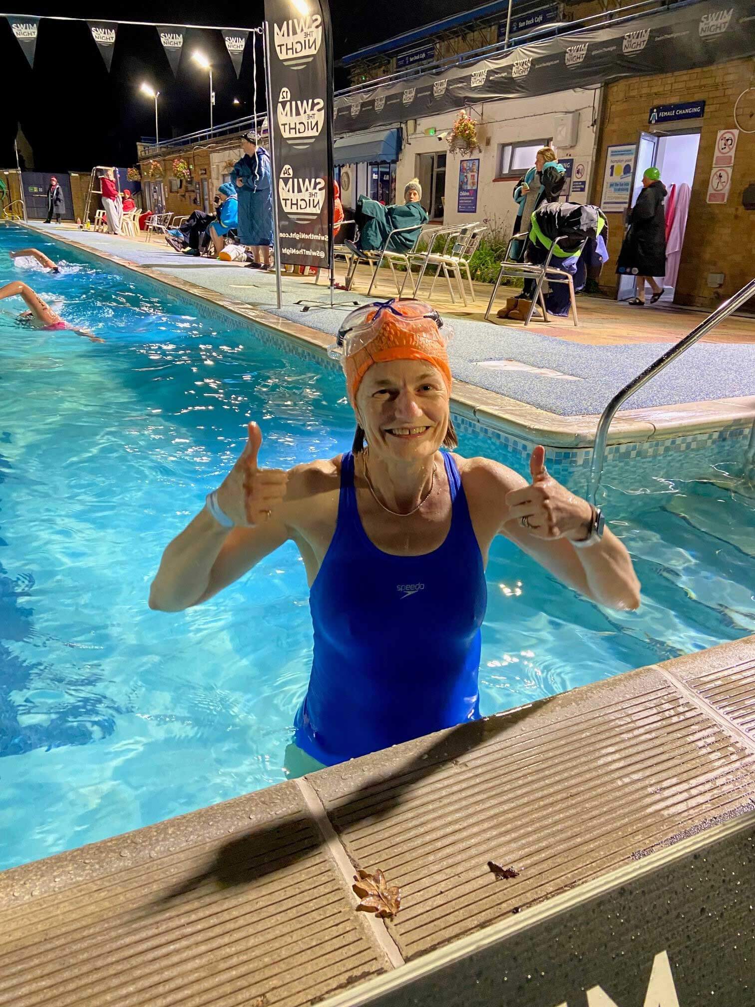 Margaret, standing & giving the thumbs up sign, in a swimming pool wearing a blue swimsuit, orange swim hat and goggles on her head, behind her is the pool with people swimming and some sitting on the side on chairs.  A banner says Swim the Night behind her