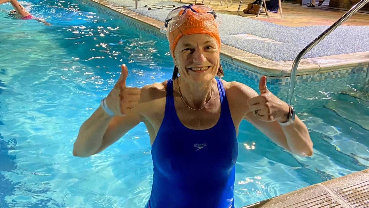 Margaret, standing & giving the thumbs up sign, in a swimming pool wearing a blue swimsuit, orange swim hat and goggles on her head, behind her is the pool with people swimming and some sitting on the side on chairs. A banner says Swim the Night behind her