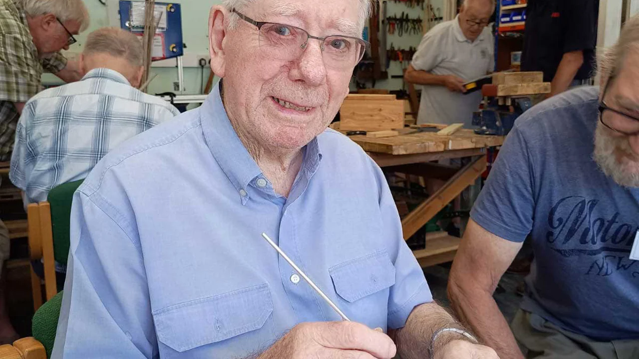 An older man wearing a blue shirt seated in a workshop holding a long thin metal rod with a wooden end. Seated next to him is a bearded man with other men working in the background at Corsham Men's Shed