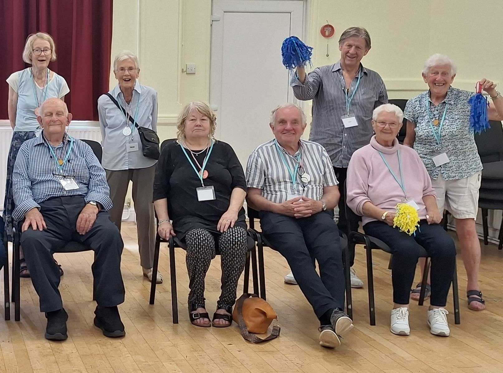 Four people sitting on chairs and four people standing behind them some waving blue and yellow pom poms at Amesbury Movement for the Mind 