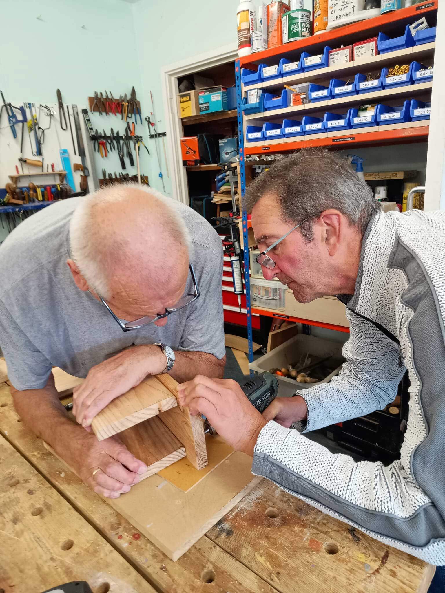 Two mean leaning over a wood construction, one is drilling and the other holding the wood both concentrating at Corsham Men's shed 